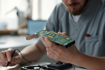 Poster - Hand of young repairman in uniform holding motherboard while sitting by workplace in repair service center and checking hardware