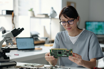 Poster - Young female technician in grey uniform looking at computer motherboard while sitting by workplace in repair service center