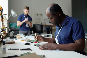 Poster - Experienced African American engineer of repair service office sitting by workplace in coworking space and checking computer motherboard