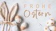 Frohe Ostern Konzept Feiertag Grußkarte mit deutschem Text - Geschenkbeutel aus Stoff mit Osterhasenohren und Ostereier auf weissem Tisch, Draufsicht