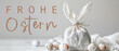 Frohe Ostern Konzept Feiertag Grußkarte mit deutschem Text - Geschenkbeutel aus Stoff mit Osterhasenohren und Ostereier auf weissem Tisch