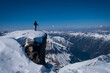 Man walking along steep mountain cliff in snow in winter, looking at mountain panorama on a sunny blue sky day.