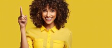 A Lively, Cheeky Woman With A Curly Long Haircut Is Wearing A Yellow Shirt And Holding Up A Peace Sign. She Looks Enthusiastic, Sassy, And Is Smiling Broadly, Winking At The Viewer.