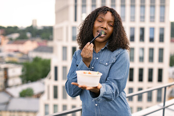 Portrait of a positive hungry African American woman holding a bowl of fresh salad in one hand and biting a slice of cucumber on fork in the other. Healthy eating concept.