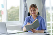 © amnaj - Smiling nurse reading from a medical chart at her desk. Healthcare and medical record-keeping concept