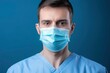© FoxTok - Portrait of young male doctor in medical clothes and mask on his face looks intently at the camera. Solid blue background. Medicine, healthcare, help and treatment of people