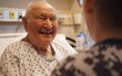 An elderly man's face lights up with a joyful smile as he interacts with a visitor in a hospital room, his eyes reflecting contentment and comfort despite the clinical surroundings.