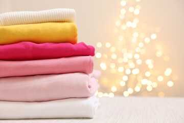 Stack of folded clothes on wooden table against beige background with blurred lights, space for text