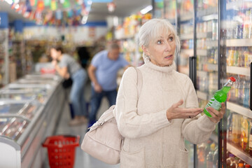 positive casual aged woman in knitted dress walking down aisle of grocery store, choosing refreshing