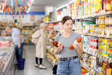 Wall Mural - Young woman choosing hot chili sauce for herself in supermarket