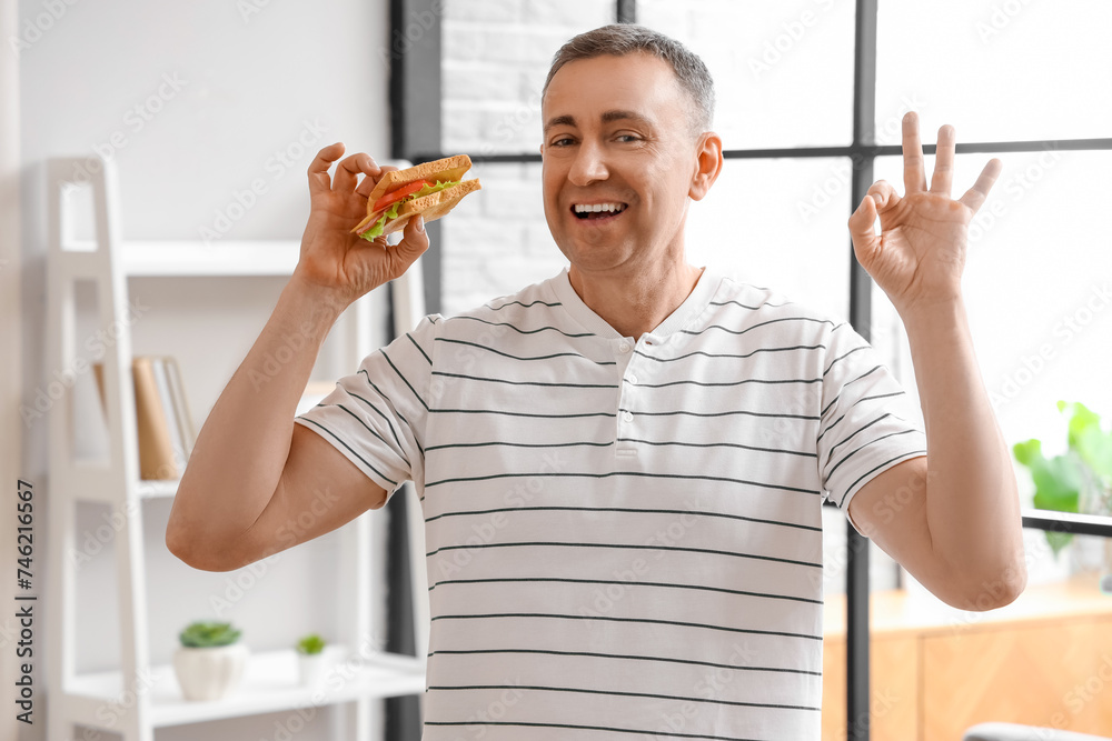Middle-aged man with delicious sandwich made from crispy toasts showing OK in kitchen
