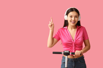 Beautiful young Asian woman in headphones with modern electric kick scooter pointing at something on pink background