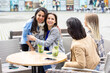 This inviting photograph captures a warm moment between friends at a city cafe. Two women are seen embracing, with one arm around the other, symbolizing a close bond. The group is engaged in a relaxed