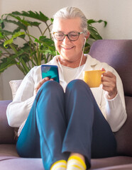 Sticker - Attractive senior grey haired woman sitting on cough at home with a cup of tea looking at mobile phone. Happy retirement lifestyle, elderly lady enjoys tech and social