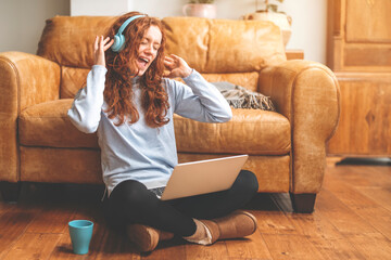 Wall Mural - Happy young redhead woman in brown shirt and using a mobile phone, listening to favorite songs, audiobooks, podcasts on headphones while sitting near a couch at home