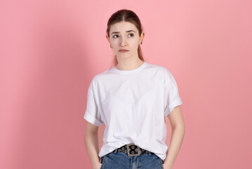 Wall Mural - Portrait of a sad and depressed attractive young brunette woman wearing a casual white t-shirt, sulking and frowning isolated on a pink studio background.