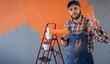 Adult bearded painter man in blue overalls over orange wall background celebrating win showing sign of victory