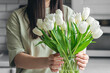 Woman putting bouquet of white tulip flowers into vase in the kitchen.