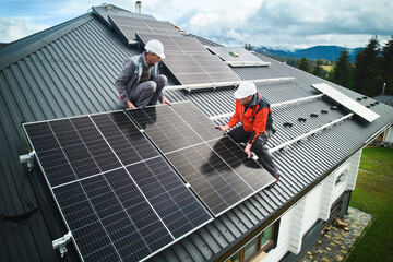 Wall Mural - Workers building photovoltaic solar module station on roof of house. Men electricians in helmets installing solar panel system outdoors. Concept of alternative and renewable energy. Aerial view.