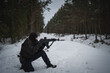 A male border guard sitting on his knee takes aim from an AK 12 assault rifle in the forest in winter, side view.