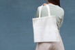 Blank white tote bag canvas fabric with handle mock up design. Back view of woman holding eco or reusable shopping bag on her sholder against blue metal wall. No plastic bag and ecology concept.