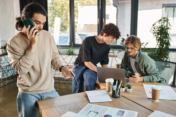 Wall Mural - Young man having phone call and looking at charts near blurred coworkers brainstorming over laptop