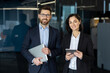 © Liubomir - Smiling male and female in stylish suits standing together in office hall and holding digital gadgets in hands. Friendly coworkers getting ready for online meeting with boss next to boardroom