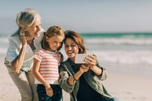 Family Beach Day Selfie With Grandmother, Mother, And Daughter Smiling By The Sea
