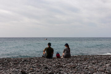 Wall Mural - Family with small child sitting on idyllic volcanic black stone beach of Praia Garajai, Canico, Madeira island, Portugal, Europe. Waves hitting shoreline of majestic Atlantic Ocean. Vacation concept