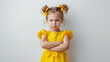 A young girl in a bright yellow dress with matching bows stands with her arms crossed, exhibiting a strong expression of displeasure and determination.
