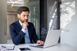 © Tetiana - A serious and thoughtful young man businessman is sitting in the office at the desk and is concentrating on his laptop