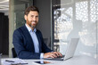 © Tetiana - Portrait of a young successful businessman, executive director in a suit working in the office at the table on a laptop, looking and smiling at the camera