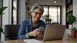 © SHERAZI - Smiling mature Hispanic woman engaged in a video call on her smartphone while working with a laptop in a bright home office Mother's day Women's day