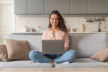 Poster - Smiling woman freelancer working on laptop while sitting on sofa at home