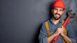 Smiling young plumber providing handyman services, holding a large red wrench, wearing a helmet, looking at the camera and smiling. Repairman with professional equipment, male worker or employee