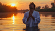 A young man wearing a white robe, standing in the river water after the baptism, praying with clasped hands and closed eyes, looking up to the sky. Sunset time. Male Christian person religious ritual