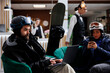Two visitors sit comfortably using digital devices while snowboard equipment suggests nearby winter sports. Image showing man and woman surfing on laptop and mobile phone in ski hotel lobby.