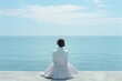 A woman in a white dress sitting peacefully by the calm sea, gazing at the clear blue sky and still water. She looks relaxed and content in the serene setting, appreciating the beauty of nature.
