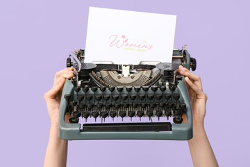 Wall Mural - Woman holding retro typewriter with greeting card for International Women's Day on lilac background, closeup