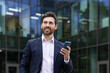 © Tetiana - Confident businessman smiling with smartphone outside modern office building