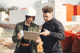 Fototapeta Tulipany - Stylish woman architect with tablet  and contractor man checking blueprints at construction site. Young engineer or construction workers in hardhat looking at plans of new modern house