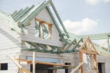 Unfinished modern farmhouse building. Wooden roof framing of mansard with dormer and aerated concrete block walls. Timber trusses, rafters and beams on blocks. New house construction