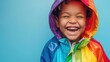 A young child with a joyful expression wearing a vibrant multicolored raincoat laughing heartily against a blue background.
