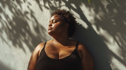 Wall Mural - A woman with curly hair wearing a black tank top leaning against a wall with her eyes closed basking in the sunlight filtering through leaves.