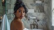 © Z A C - A woman with dark hair styled in an updo wearing a white robe standing in a luxurious bathroom with marble walls and a chandelier.