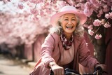 Fototapeta  - happy smiling elderly woman riding a bicycle through a park with pink cherry blossoms