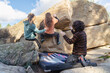 © ADDICTIVE STOCK - Back view of three friends engage in bouldering on a large rock, with one spotting for safety, against a backdrop of clear skies