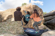 © ADDICTIVE STOCK - Back view of three friends engage in bouldering on a large rock, with one spotting for safety, against a backdrop of clear skies