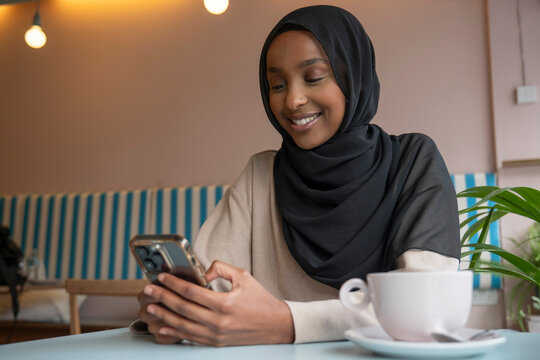 Smiling young woman in hijab using phone in cafe