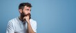 © Koja - A bearded man looks lost in thought with his chin resting on his hand, against a blue background. An imaginary look that gets lost in the distance. Pastel blue background.